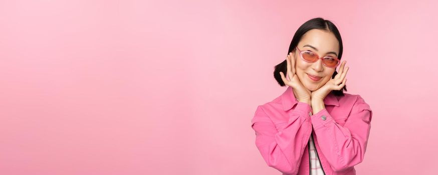 Portrait of stylish cute asian girl, smiling and touching face, looking up dreamy, thoughtful look, standing over pink background.