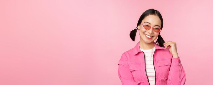 Beautiful asian girl in sunlgasses, smiling at camera, posing against pink studio background.