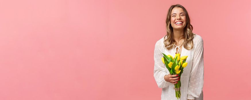 Holidays, beauty and spring concept. Carefree happy attractive blond girl in white dress holding yellow tulips and laughing with pleased expression, standing pink background, have romantic date.