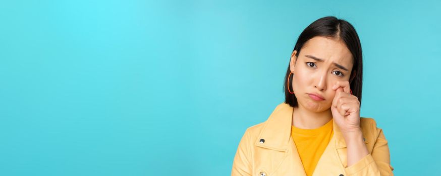 Close up of sad asian girl crying, wiping tears and sulking, looking upset and gloomy, posing against blue background. Copy space
