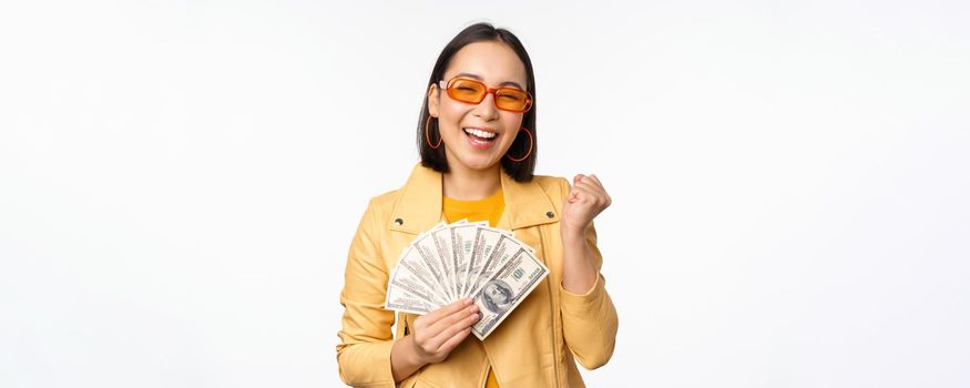 Stylish smiling asian girl holding money cash, showing dollars and celebrating, standing over white background. Copy space
