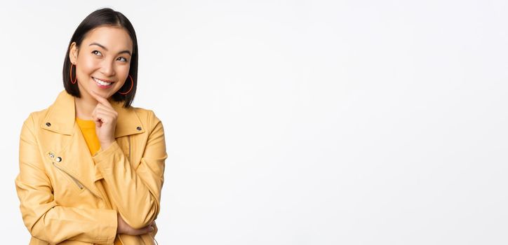 Portrait of stylish korean woman in yellow jacket, smiling thoughtful, thinking and looking up at logo or advertisement, standing over white background.