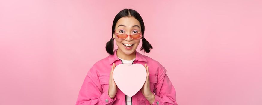 Beautiful asian girl smiling happy, showing heart gift box and looking excited at camera, standing over pink romantic background.