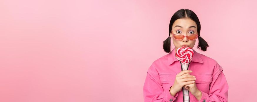 Silly and cute asian female model licking lolipop, eating candy sweet and smiling, looking excited, standing over pink background.