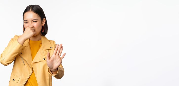 Image of asian girl shuts nose and reject smth with disgusting bad smell, shuts nose, standing over white background. Copy space