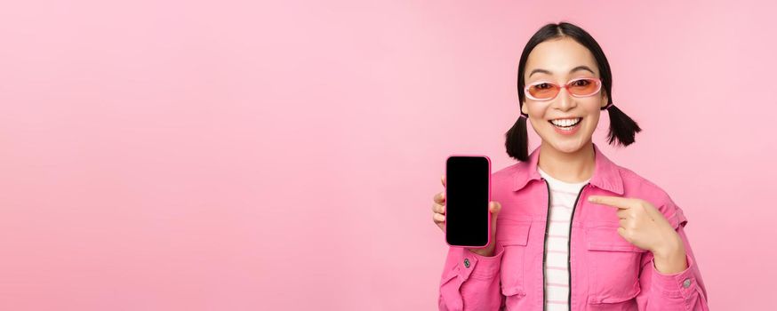 Enthusiastic asian woman in stylish clothes, sunglasses, pointing finger at mobile phone screen, showing smartphone application, standing over pink background.