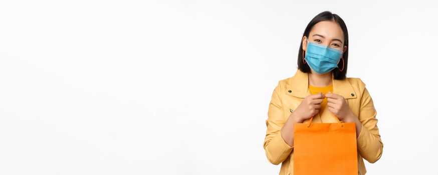 Covid-19 and shop concept. Young asian stylish woman, wearing medical face mask, holding shopping bag, going in malls during pandemic, white background.