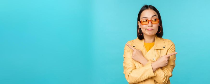 Indecisive asian woman in sunglasses, makes choice between two variants, points sideways, looks puzzled, stands over blue background.