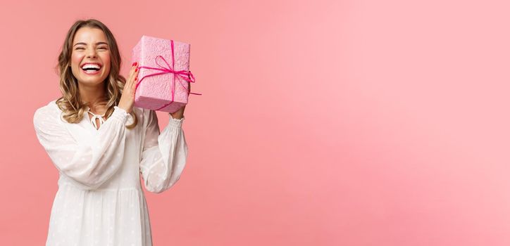 Holidays, celebration and women concept. Portrait of happy charismatic blond girl shaking gift box wondering whats inside as celebrating birthday, receive b-day presents, pink background.