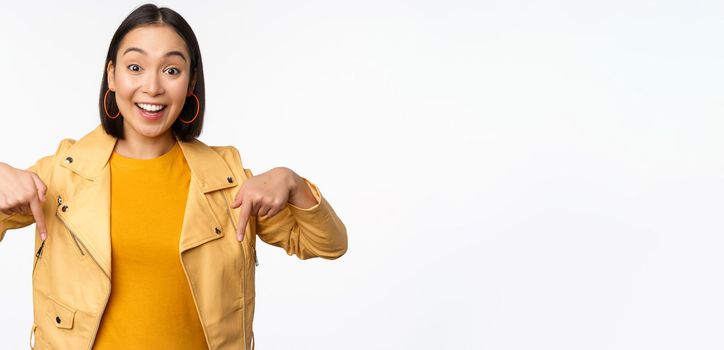 Portrait of happy smiling asian girl, pointing fingers down and showing logo, demonstrating banner, standing in yellow jacket against white background. Copy space