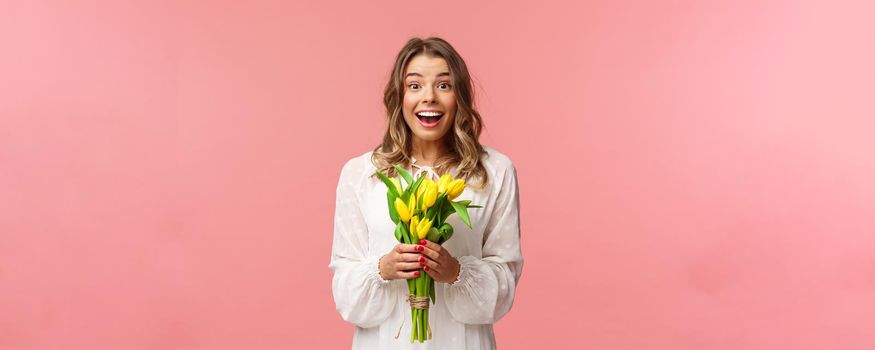 Holidays, beauty and spring concept. Portrait of excited and amazed young beautiful blond girl in white dress, holding yellow tulips, receive flowers as gift, smling amused, pink background.