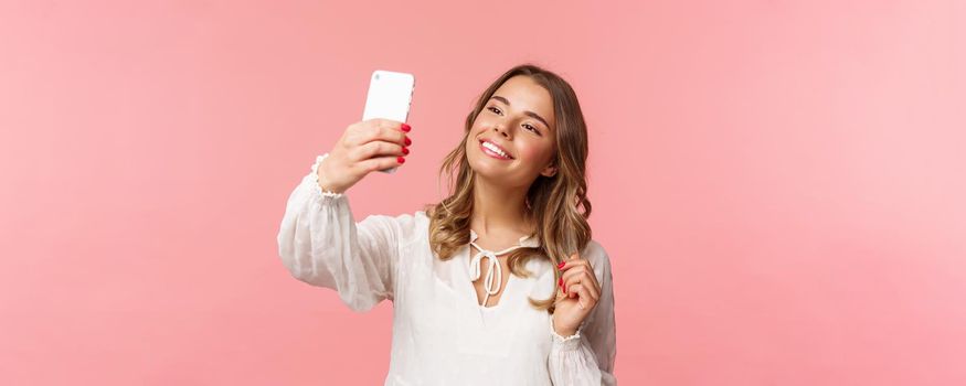 Close-up portrait of beautiful and stylish blond girl taking selfie in park at spring, gather likes and followers, make blogger content, taking photo on smartphone, smiling sassy, pink background.