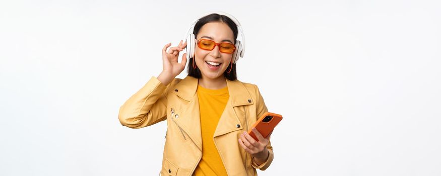 Happy stylish asian woman in sunglasses, listening music in headphones, holding smartphone, singing and dancing, standing over white background.