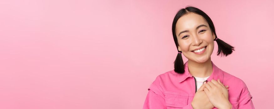 Cute korean girl with silly hairbuns, looking happy and grateful, thank you pose, holding hands on heart flattered, posing against pink background.