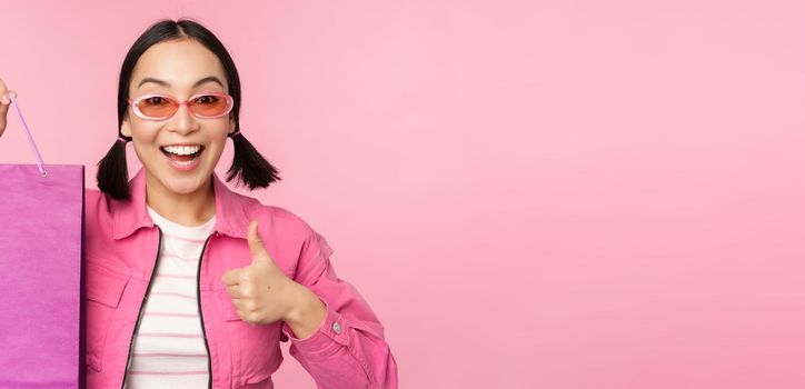 Shopping. Stylish asian girl in sunglasses, showing bag from shop and smiling, recommending sale promo in store, standing over pink background.
