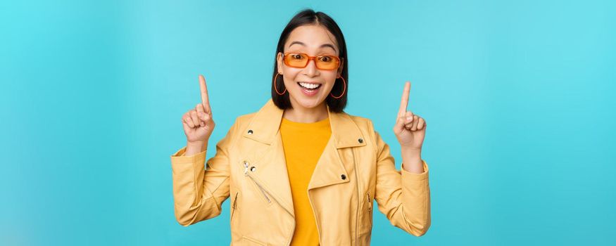 Enthusiastic asian girl in sunglasses, points fingers up, shows banner or logo on top, stands over blue background. Copy space