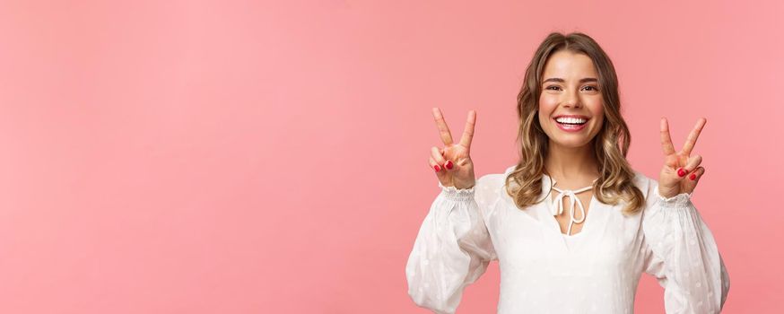 Close-up portrait of optimistic kawaii young blond girl with lovely beaming smile, showing peace signs and looking camera with positive attitude, enjoying spring, white dress, pink background.
