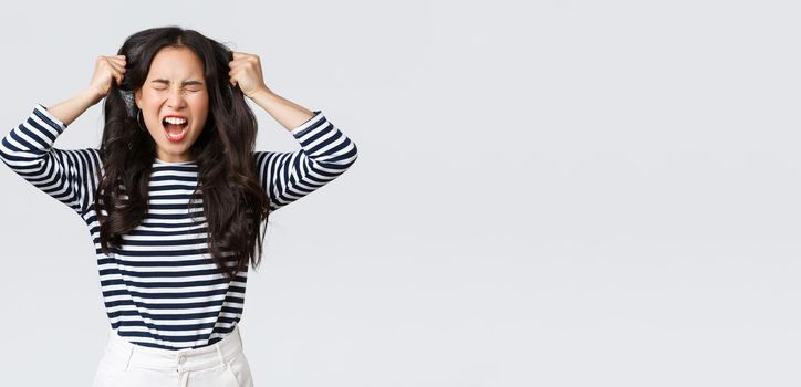 Lifestyle, people emotions and casual concept. Pissed-off mad and angry asian young woman tossing hair, pulling it from head with screams and closed eyes, standing bothered white background.