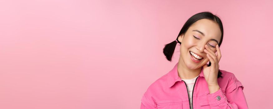 Close up portrait of beautiful asian girl looking enthusiastic and smiling, laughing and smiling, standing happy against pink background.