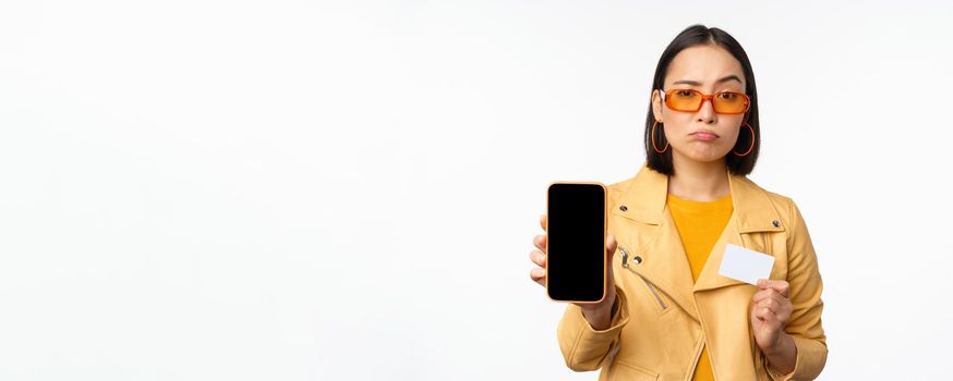 Sad asian girl in sunglasses, showing smartphone app interface, credit card, looking disappointed, standing over white background. Copy space