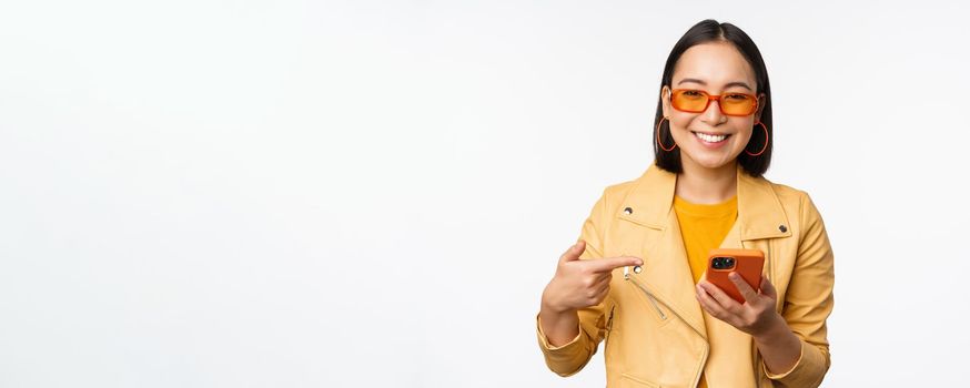 Beautiful smiling asian girl in sunglasses, pointing finger at smartphone, showing app, store on mobile phone, standing over white background.