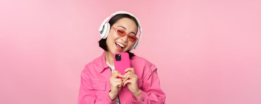 Stylish asian girl in headphones, listening music and taking photos on mobile phone, using smartphone, standing over pink background.