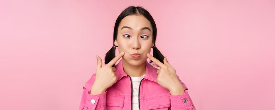 Close up portrait of young asian girl showing her dimples, poking cheeks silly and making funny faces, standing over pink background.