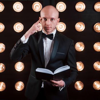 He know what you thinking about. Magician in black suit standing in the room with special lighting at backstage.