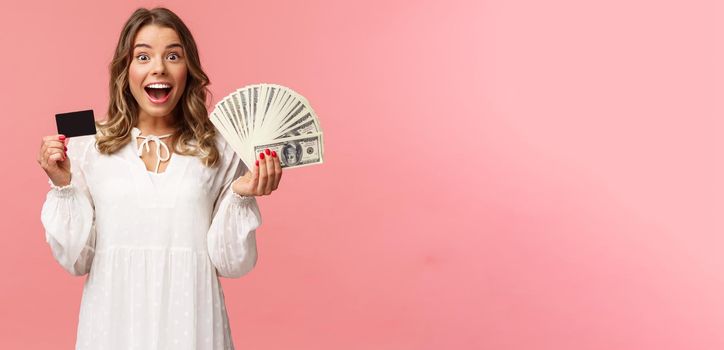 Portrait of excited happy good-looking blond girl in white dress, winning money, placed good bet, made deal, holding dollars money and credit card, smiling amused at camera, pink background.