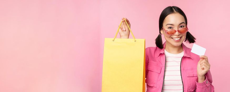 Happy young asian woman shows credit card and shopping bag, store sale announcement, buying smth in shop, posing against pink background.