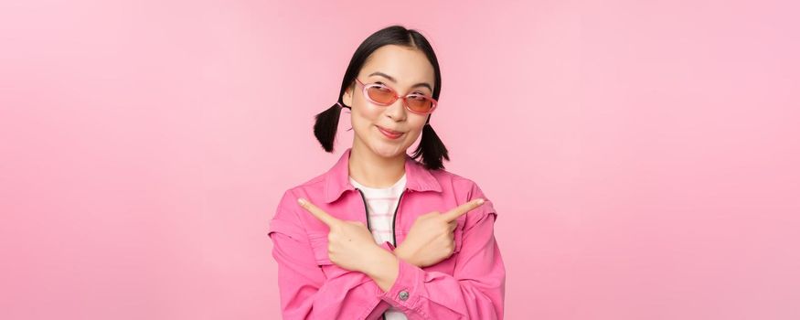 Choice. Stylish korean girl, asian female model points fingers sideways, shows two variants, product advertisement, demonstrating items, standing over pink background.