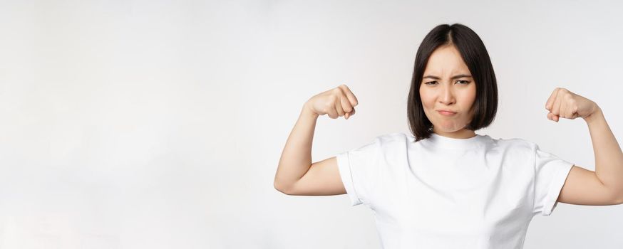 Smiling asian woman showing flexing biceps, muscles strong arms gesture, standing in white tshirt over white background.