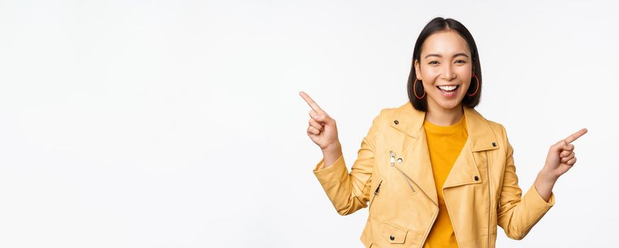 Happy beautiful asian girl pointing fingers left and right, showing banner logo, demonstrating sale, standing in yellow jacket over white background.