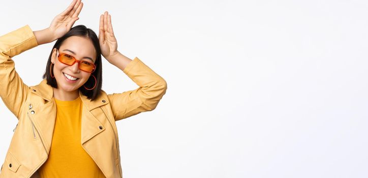 Portrait of beautiful asian girl in sunglasses, showing bunny ears hands gesture and smiling, standing happy over white background.