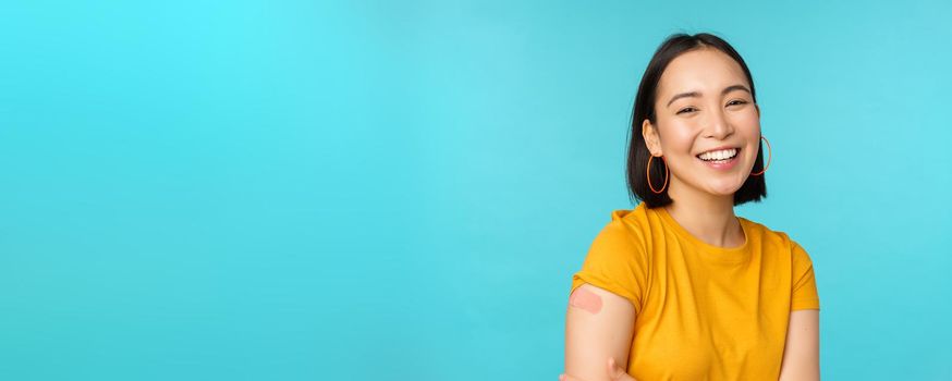 Vaccine campaign from covid-19. Happy and healthy asian girl laughing after vaccination from coronavirus, bandaid on shoulder, wearing yellow t-shirt, blue background.