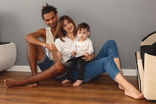 Portrait of happy, modern, multiracial family with a little boy in casual clothes sits against a gray wall in a room, looks at camera, smiling. copy space