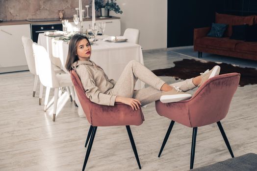 photo of a women in beige sports suit and stylish leather sneakers posing in the chair on a kitchen . selective focus. Indoors