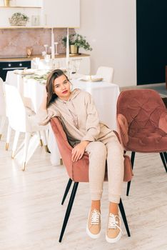 photo of a women in beige sports suit and stylish leather sneakers posing in the chair on a kitchen . selective focus. Indoors