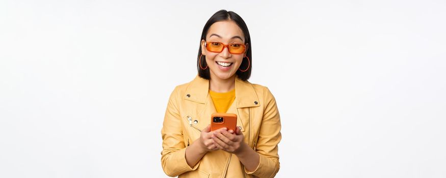 Modern asian girl in sunglasses using her mobile phone, smiling and looking happy, posing against white background. Copy space