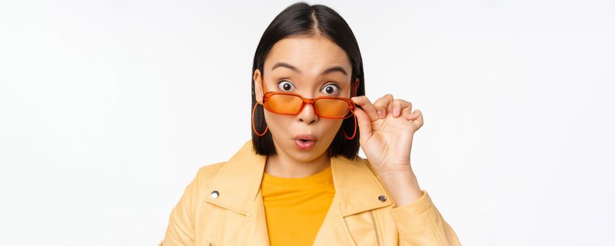 Close up portrait of asian girl looking surprised, wow face, takes off sunglasses and staring impressed at camera, standing over white background.