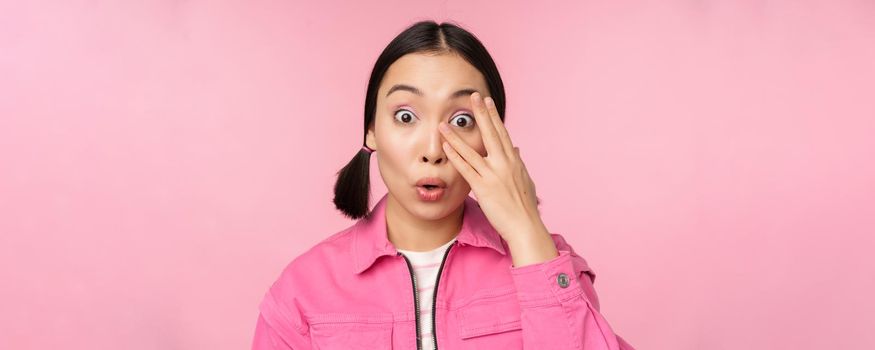 Close up portrait of young asian girl looking surprised, express amazement and wonder, peeking through fingers, standing over pink background.