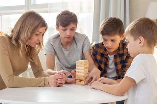 A family, three boys and a woman, are enthusiastically playing a board game made of wooden rectangular blocks stacked in the form of a tower in a sunny room. Close-up