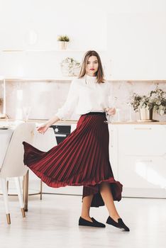 Portrait of fashionable woman in a red skirt, white blouse and stylish suede shoes with a buckle posing on the kitchen. Girl with a big red lips