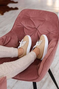 photo of a women legs in beige sports suit and stylish leather sneakers posing in the chair. selective focus. Indoors