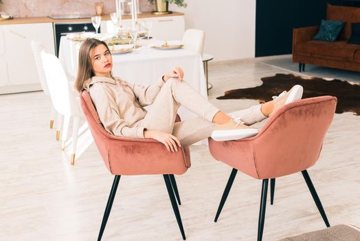 photo of a women in beige sports suit and stylish leather sneakers posing in the chair on a kitchen . selective focus. Indoors