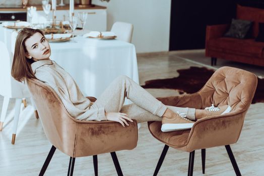 photo of a women in beige sports suit and stylish leather sneakers posing in the chair on a kitchen . selective focus. Indoors