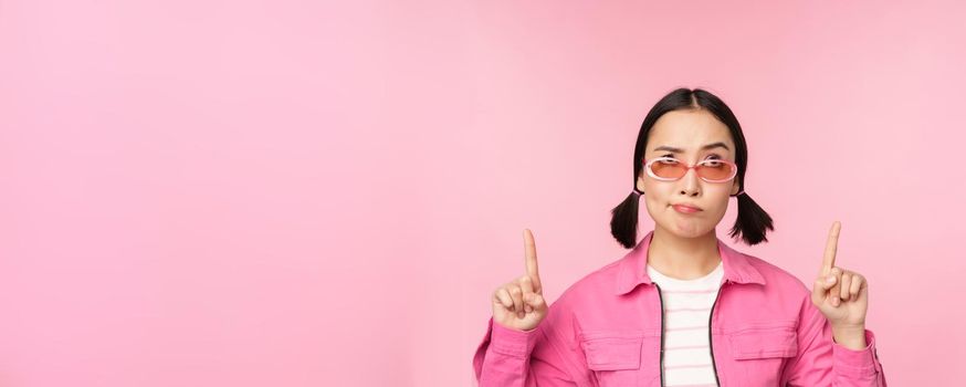 Hesitant asian girl, wears sunglasses, frowns and points fingers up, looks with disbelief, skeptical face expression, stands over pink background.