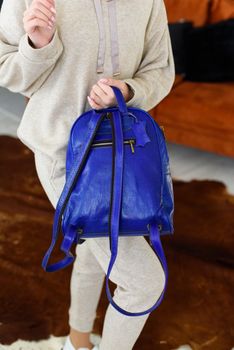 photo of a blue leather backpack in womens hand. Girl wearing beige sports suit. selective focus. Indoors