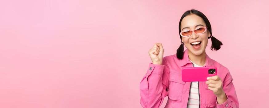 Happy smiling korean girl winning on mobile phone, looking at horizontal smartphone screen and rejoicing, achieve goal, celebrating, standing over pink background.