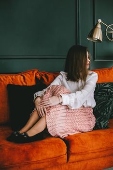 Portrait of fashionable young woman in a beige skirt, white blouse and stylish beige black seude shoes with a buckle posing on a orange sofa. Girl with a big dark red lips